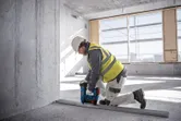 Seseorang yang mengenakan perlengkapan keselamatan menggunakan paku beton tanpa kabel untuk mengencangkan rel logam ke lantai.