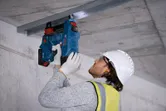 Seseorang yang mengenakan peralatan keselamatan mengamankan saluran logam ke langit-langit beton dengan paku beton tanpa kabel.
