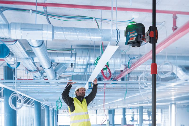 Seseorang yang mengenakan peralatan keselamatan memasang panel langit-langit menggunakan alat perata laser pada tiang teleskopik.