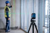 Seorang pekerja yang mengenakan perlengkapan keselamatan sedang menyelaraskan rangka dinding menggunakan alat perata laser.