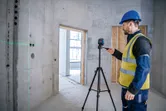 Seseorang yang mengenakan perlengkapan keselamatan mengoperasikan alat perataan laser pada tripod di ruang beton.