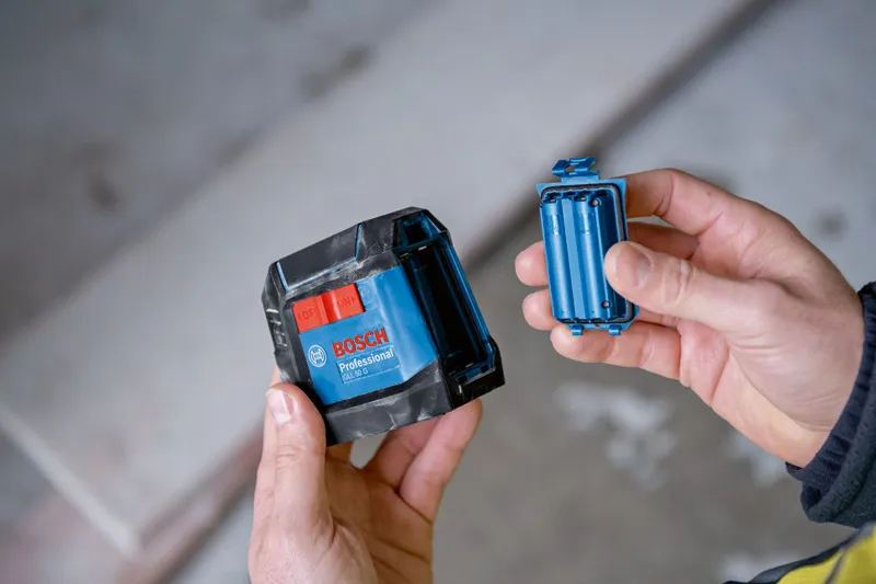 Seseorang memegang alat perataan laser biru dan kompartemen baterainya di dalam ruangan.