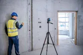 Seseorang yang mengenakan peralatan keselamatan menandai dinding beton menggunakan alat perata laser pada tripod.
