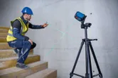 Seseorang yang mengenakan perlengkapan keselamatan menandai dinding menggunakan alat perata laser hijau pada tripod.