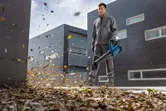 Seseorang yang mengenakan peralatan keselamatan membersihkan dedaunan dengan blower daun tanpa kabel di luar gedung.