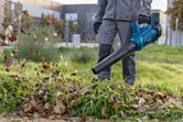 Seseorang yang mengenakan peralatan keselamatan menggunakan blower daun tanpa kabel untuk membersihkan daun di rumput.