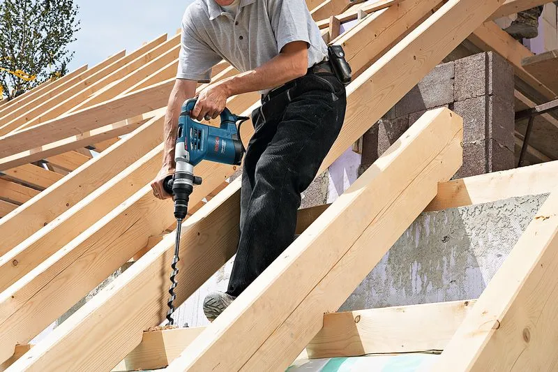 Seseorang mengebor balok kayu pada struktur atap, tidak mengenakan peralatan keselamatan.