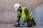 Seseorang yang mengenakan perlengkapan keselamatan memotong tulangan beton yang terbuka dengan penggiling sudut.