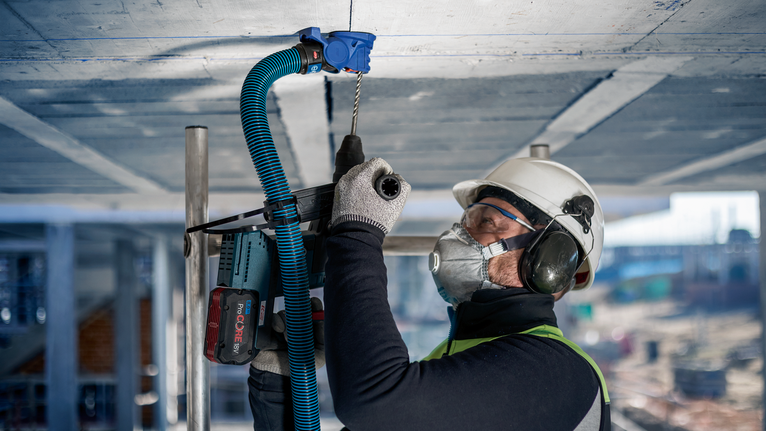 Seseorang yang mengenakan peralatan keselamatan mengebor langit-langit beton di lokasi konstruksi.
