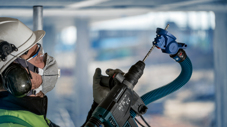 Seseorang yang mengenakan peralatan keselamatan mengebor langit-langit menggunakan alat listrik dengan penyedot debu.