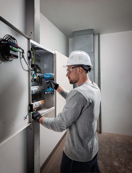 Orang yang mengenakan peralatan keselamatan menggunakan bor tanpa kabel pada panel listrik.