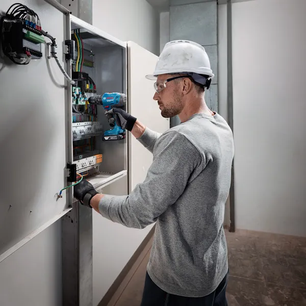 Seseorang yang mengenakan peralatan keselamatan menggunakan bor tanpa kabel pada panel listrik.