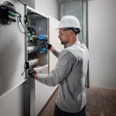 Seseorang yang mengenakan peralatan keselamatan menggunakan bor tanpa kabel pada panel listrik.