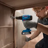 Seseorang yang mengenakan peralatan keselamatan menggunakan bor tanpa kabel untuk memasang engsel kabinet.