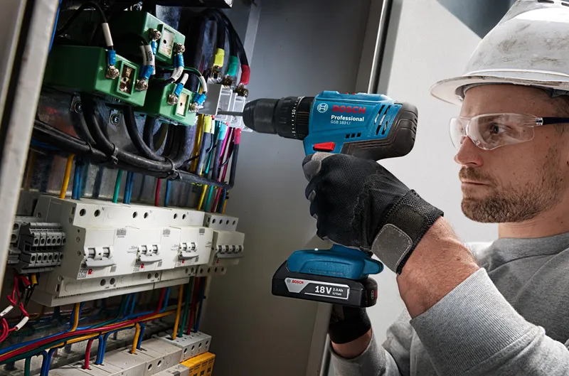 Seseorang yang mengenakan peralatan keselamatan mengebor panel kontrol yang penuh dengan kabel.