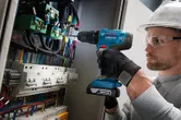 Seseorang yang mengenakan peralatan keselamatan mengebor panel kontrol yang penuh dengan kabel.