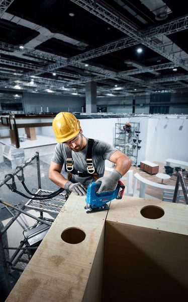 Seseorang yang mengenakan perlengkapan keselamatan menggunakan gergaji ukir untuk memotong kayu di lokasi konstruksi.
