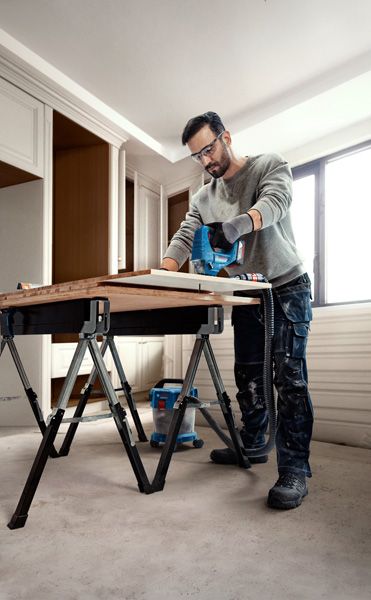 Seseorang yang mengenakan perlengkapan keselamatan menggunakan gergaji ukir untuk memotong kayu di meja kerja.