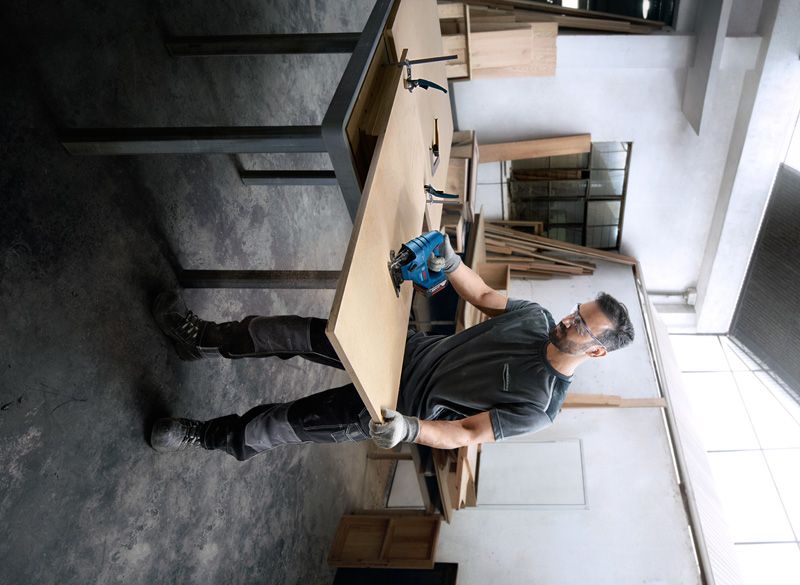 Seseorang yang mengenakan perlengkapan keselamatan memotong kayu dengan perkakas listrik di bengkel.