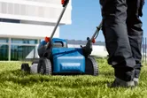 Seseorang yang mengenakan peralatan keselamatan mendorong mesin pemotong rumput tanpa kabel di atas rumput hijau.