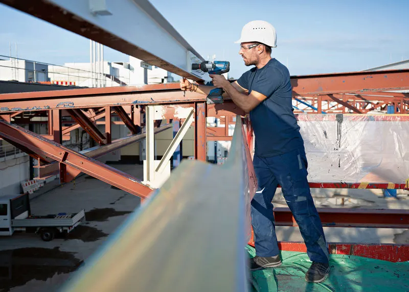 Seseorang yang mengenakan perlengkapan keselamatan menggunakan kunci pas benturan tanpa kabel pada balok baja.