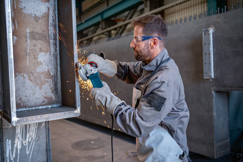 Seseorang yang mengenakan peralatan keselamatan menggiling logam dengan penggiling sudut, yang menghasilkan percikan api.