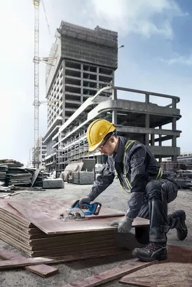 Seseorang yang mengenakan peralatan keselamatan memotong kayu lapis di lokasi konstruksi dengan gergaji tanpa kabel.