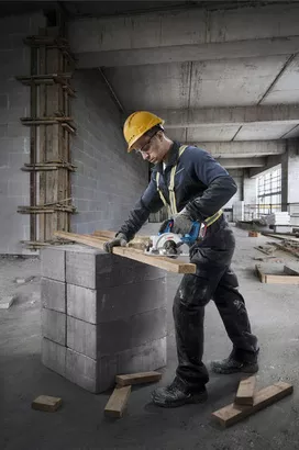 Seseorang yang mengenakan peralatan keselamatan memotong kayu dengan gergaji bundar pada balok beton.