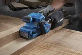 Seseorang yang mengenakan perlengkapan keselamatan mengampelas papan kayu dengan mesin pengamplas sabuk biru.