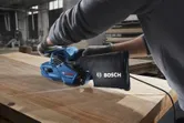 Seseorang yang mengenakan perlengkapan keselamatan mengampelas papan kayu dengan mesin pengamplas sabuk biru.
