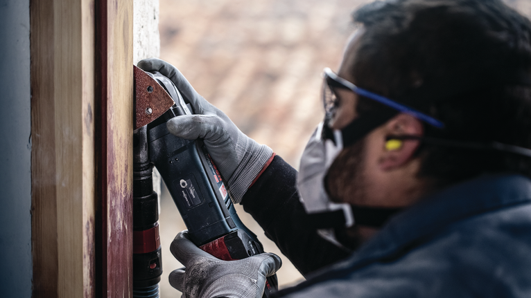 Orang yang mengenakan peralatan keselamatan mengampelas rangka kayu menggunakan perkakas listrik.