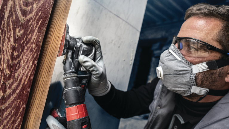 Orang yang mengenakan peralatan keselamatan mengampelas permukaan kayu dengan perkakas listrik.