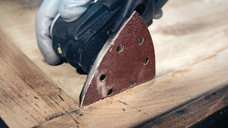 Orang yang mengenakan peralatan keselamatan mengampelas permukaan kayu dengan mesin pengamplas detail.