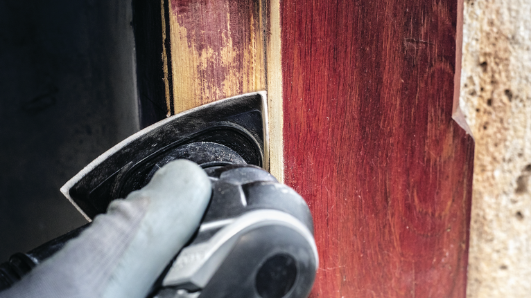 Orang yang mengenakan peralatan keselamatan mengampelas kusen pintu kayu dengan perkakas listrik.