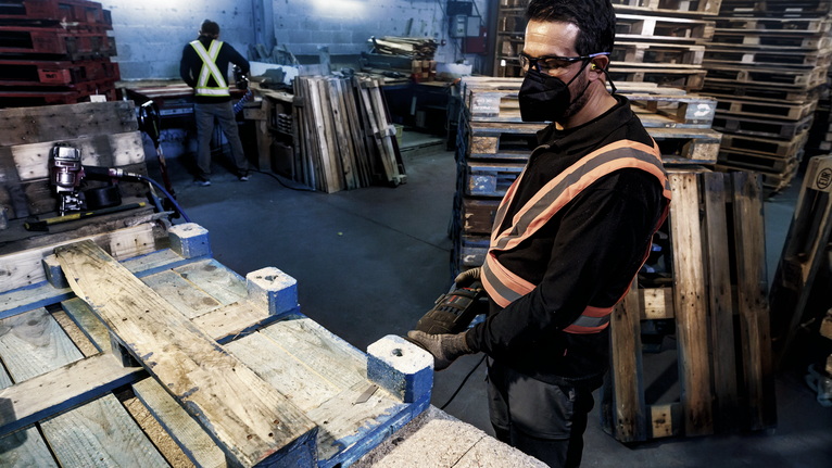 Seseorang yang mengenakan perlengkapan keselamatan menggunakan perkakas listrik untuk bekerja di atas palet kayu.