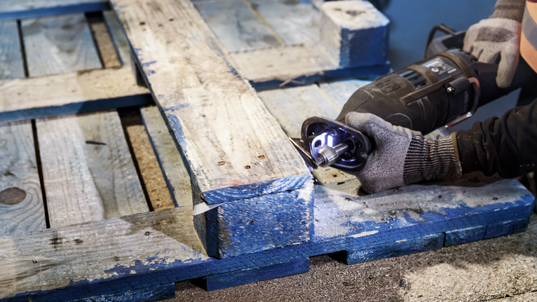 Orang yang mengenakan peralatan keselamatan memotong palet kayu dengan gergaji bolak-balik.