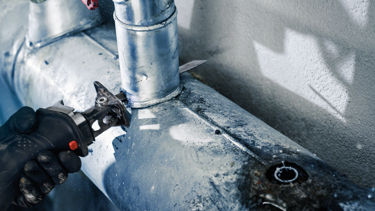 Orang yang mengenakan peralatan keselamatan memotong pipa logam dengan alat listrik.