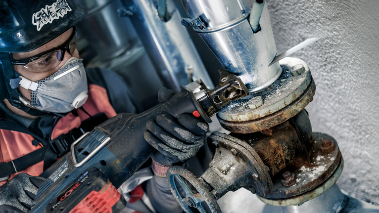 Seseorang yang mengenakan perlengkapan keselamatan menggunakan gergaji bolak-balik untuk memotong pipa yang berkarat.