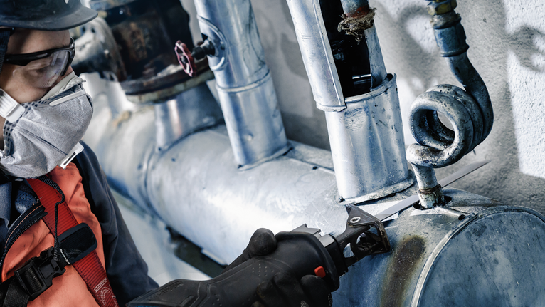 Orang yang mengenakan peralatan keselamatan memotong pipa logam dengan alat listrik.