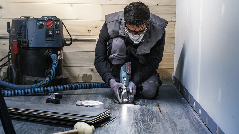 Seseorang yang mengenakan peralatan keselamatan memotong lantai laminasi dengan perkakas listrik.