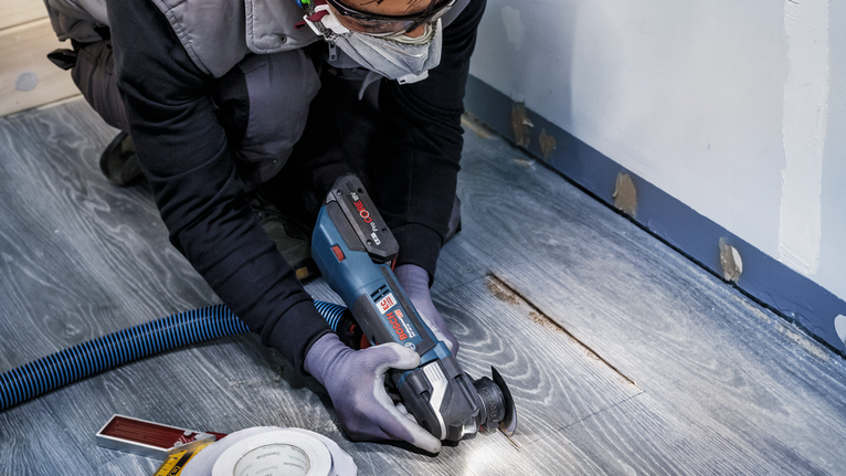 Orang yang mengenakan peralatan keselamatan memotong lantai kayu dengan alat listrik.