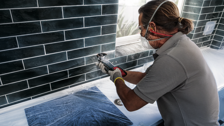 Orang yang mengenakan peralatan keselamatan menggunakan alat listrik untuk memotong ubin di bawah jendela.