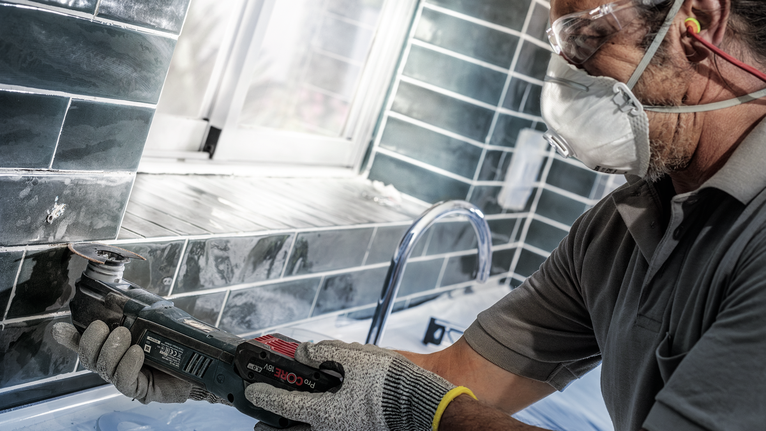 Seseorang yang mengenakan peralatan keselamatan menggunakan alat listrik untuk memotong ubin di dekat wastafel.