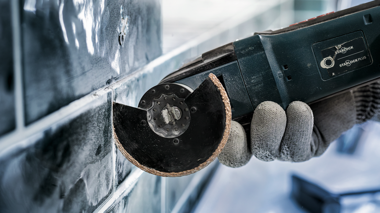 Seseorang yang mengenakan perlengkapan keselamatan menggunakan perkakas listrik untuk memotong ubin.