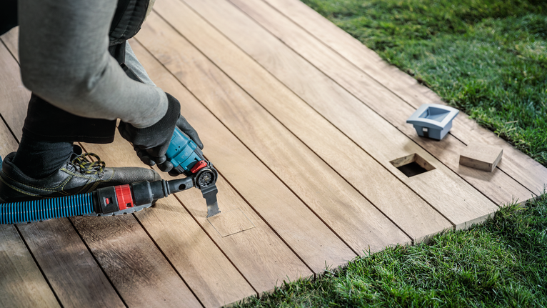 Orang yang mengenakan peralatan keselamatan memotong dek kayu dengan alat listrik.