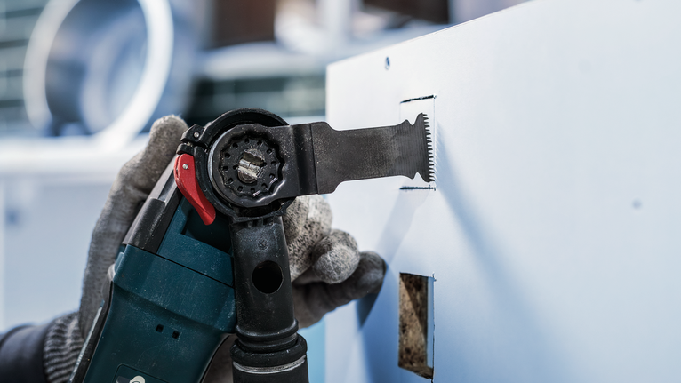 Orang yang mengenakan peralatan keselamatan memotong lubang persegi panjang di dinding kering dengan alat listrik.