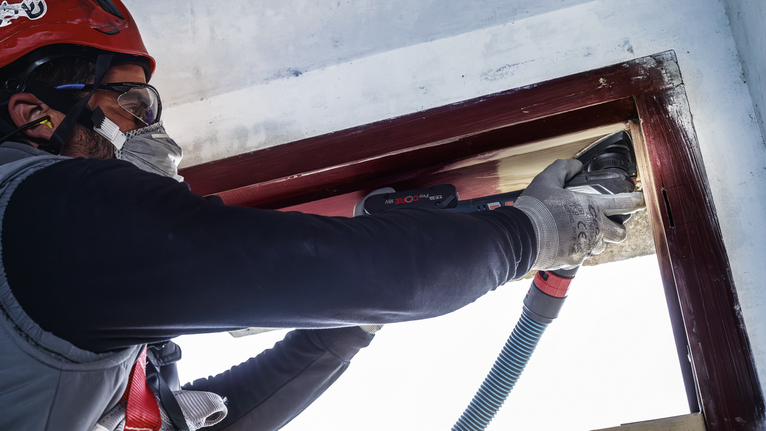 Orang yang mengenakan peralatan keselamatan sedang mengampelas bingkai jendela kayu dengan perkakas listrik.