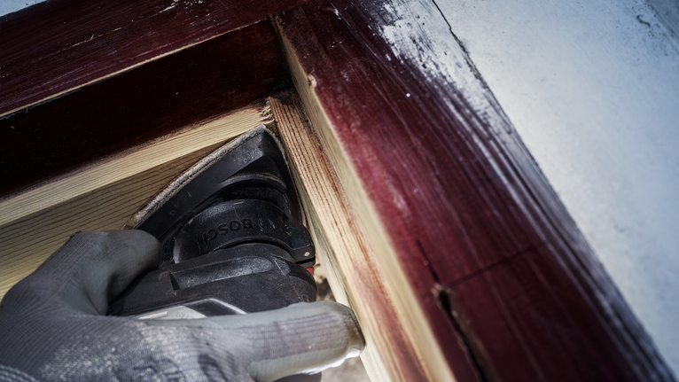 Orang yang mengenakan peralatan keselamatan mengampelas kayu di dekat bingkai jendela yang dicat.