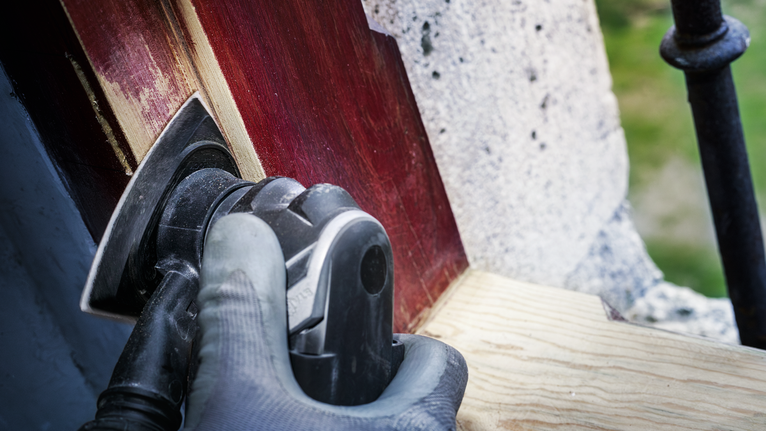 Orang yang mengenakan peralatan keselamatan sedang mengampelas kayu yang dicat dengan alat listrik.