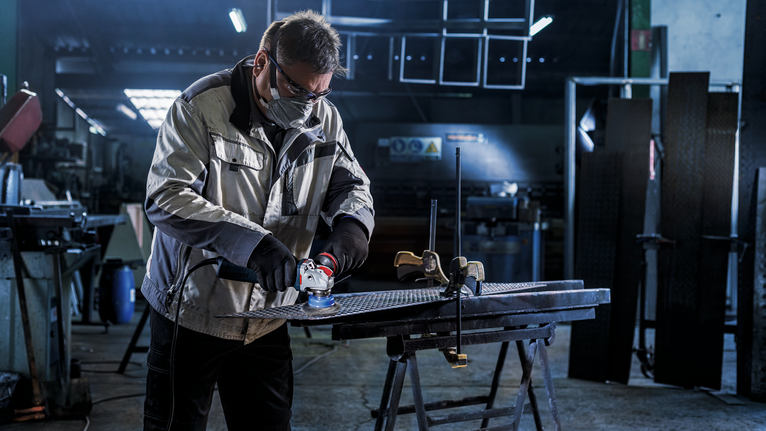 Seseorang yang mengenakan perlengkapan keselamatan sedang menggiling lembaran logam di bengkel.